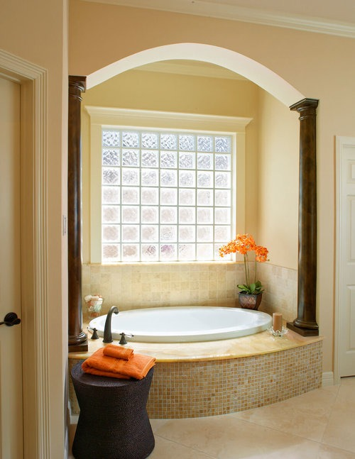 Luxurious bathroom with an arched alcove, a round soaking tub, decorative columns, and a glass-block window, creating a serene bathing area.